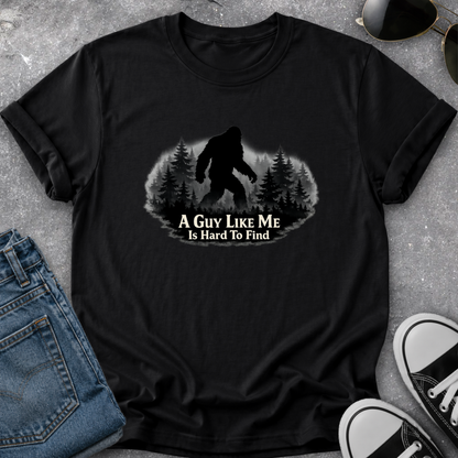 A black short-sleeve graphic t-shirt laid flat on a gray textured surface, styled with blue jeans, sunglasses, and black-and-white sneakers. The shirt features a graphic of a Bigfoot silhouette in a pine forest with the text 'A GUY LIKE ME Is Hard To Find'.