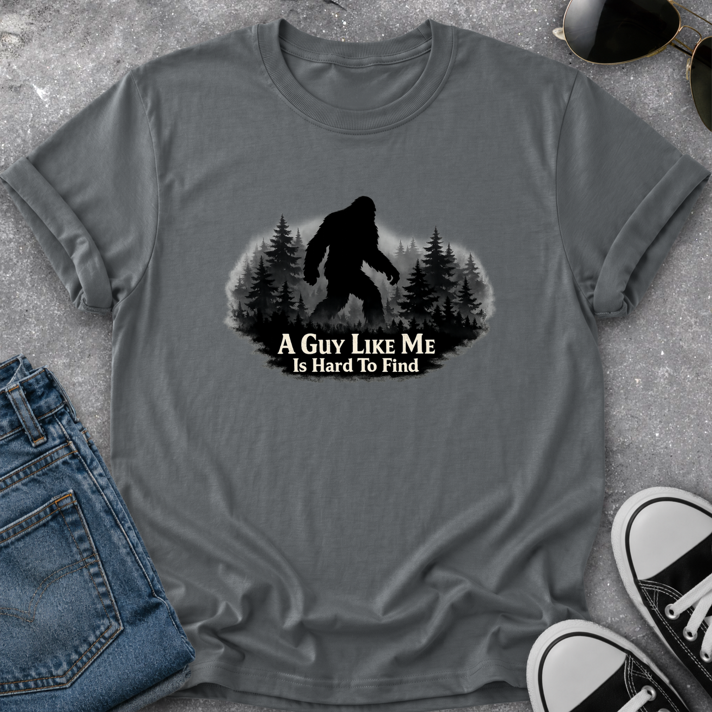A gray short-sleeve t-shirt with a black graphic of a Bigfoot silhouette in a pine forest and the text 'A GUY LIKE ME IS HARD TO FIND'. The shirt is styled in a flat-lay with blue jeans, sunglasses, and black-and-white sneakers on a gray textured surface.