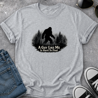 A heather gray short-sleeve t-shirt laid flat on a textured gray surface, featuring a black graphic of a Bigfoot silhouette among pine trees with the text 'A GUY LIKE ME IS HARD TO FIND.' Blue jeans, sunglasses, and black-and-white sneakers are partially visible around the shirt as styling props.