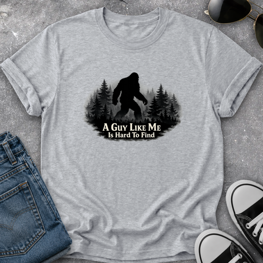 A heather gray short-sleeve t-shirt laid flat on a textured gray surface, featuring a black graphic of a Bigfoot silhouette among pine trees with the text 'A GUY LIKE ME IS HARD TO FIND.' Blue jeans, sunglasses, and black-and-white sneakers are partially visible around the shirt as styling props.