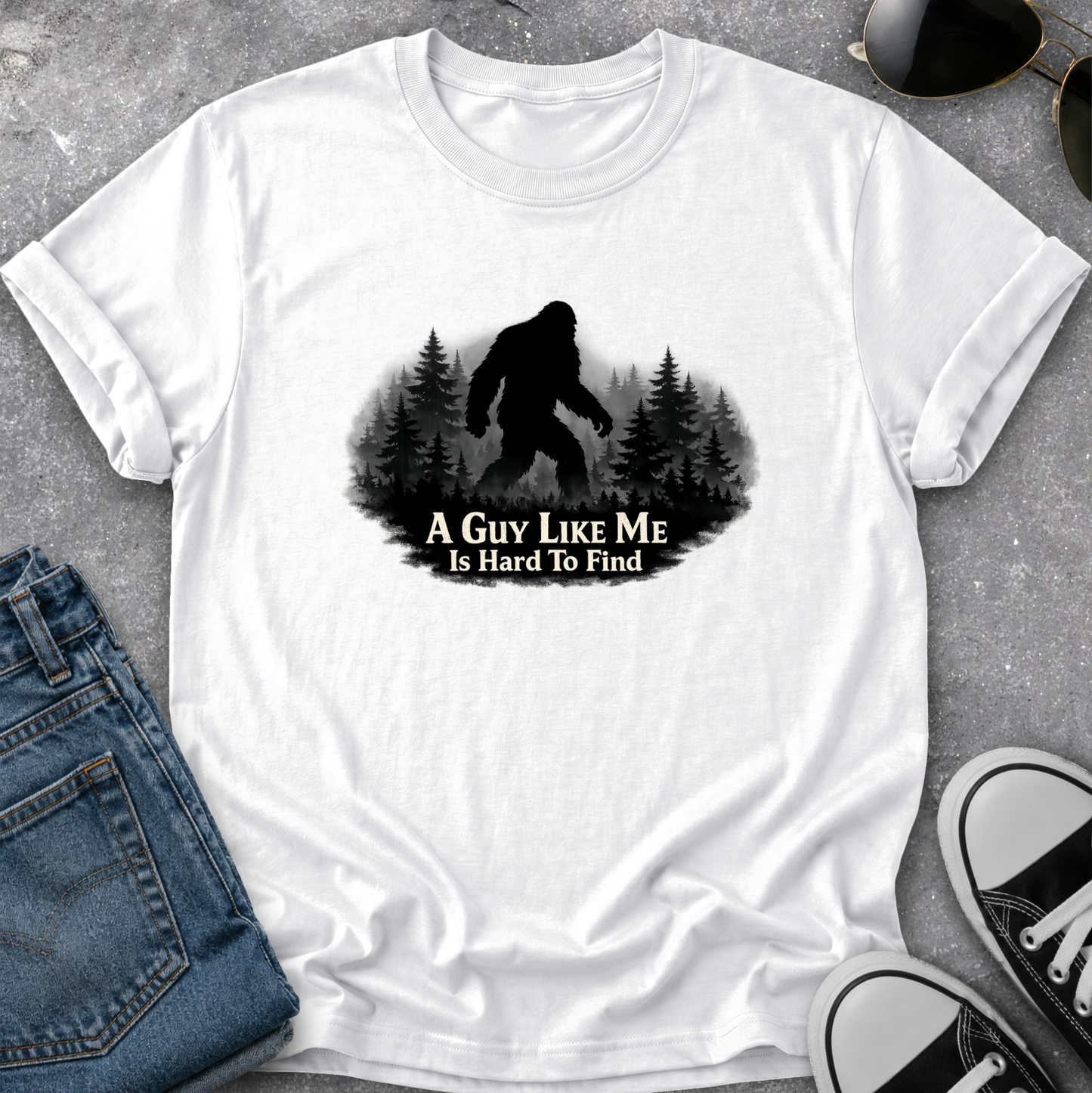 A white short-sleeve graphic T-shirt laid flat on a gray textured surface, styled with blue jeans, sunglasses, and black-and-white sneakers. The shirt has a black silhouette of a Bigfoot figure in a forest with the text 'A GUY LIKE ME IS HARD TO FIND' printed on the front.