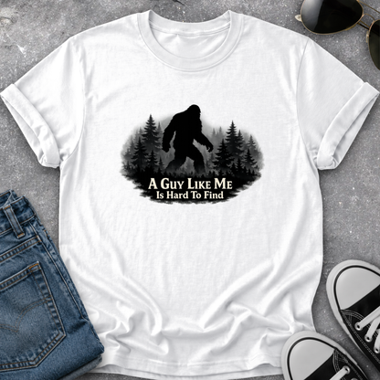 A white short-sleeve graphic T-shirt laid flat on a gray textured surface, styled with blue jeans, sunglasses, and black-and-white sneakers. The shirt has a black silhouette of a Bigfoot figure in a forest with the text 'A GUY LIKE ME IS HARD TO FIND' printed on the front.