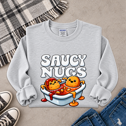 A light gray crewneck sweatshirt with a graphic of two cartoon chicken nuggets in a bowl of sauce under the text 'SAUCY NUGS'. The sweatshirt is styled in a flat lay with a plaid blanket, sneakers, and jeans on a concrete background.