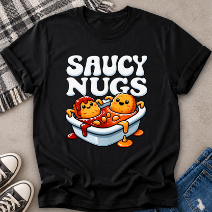 A black short-sleeve graphic T-shirt laid flat on a light gray surface with a plaid blanket, sneakers, and jeans partially visible around it. The shirt features large white 'SAUCY NUGS' text and a cartoon graphic of two orange nuggets in a white bowl with red sauce.