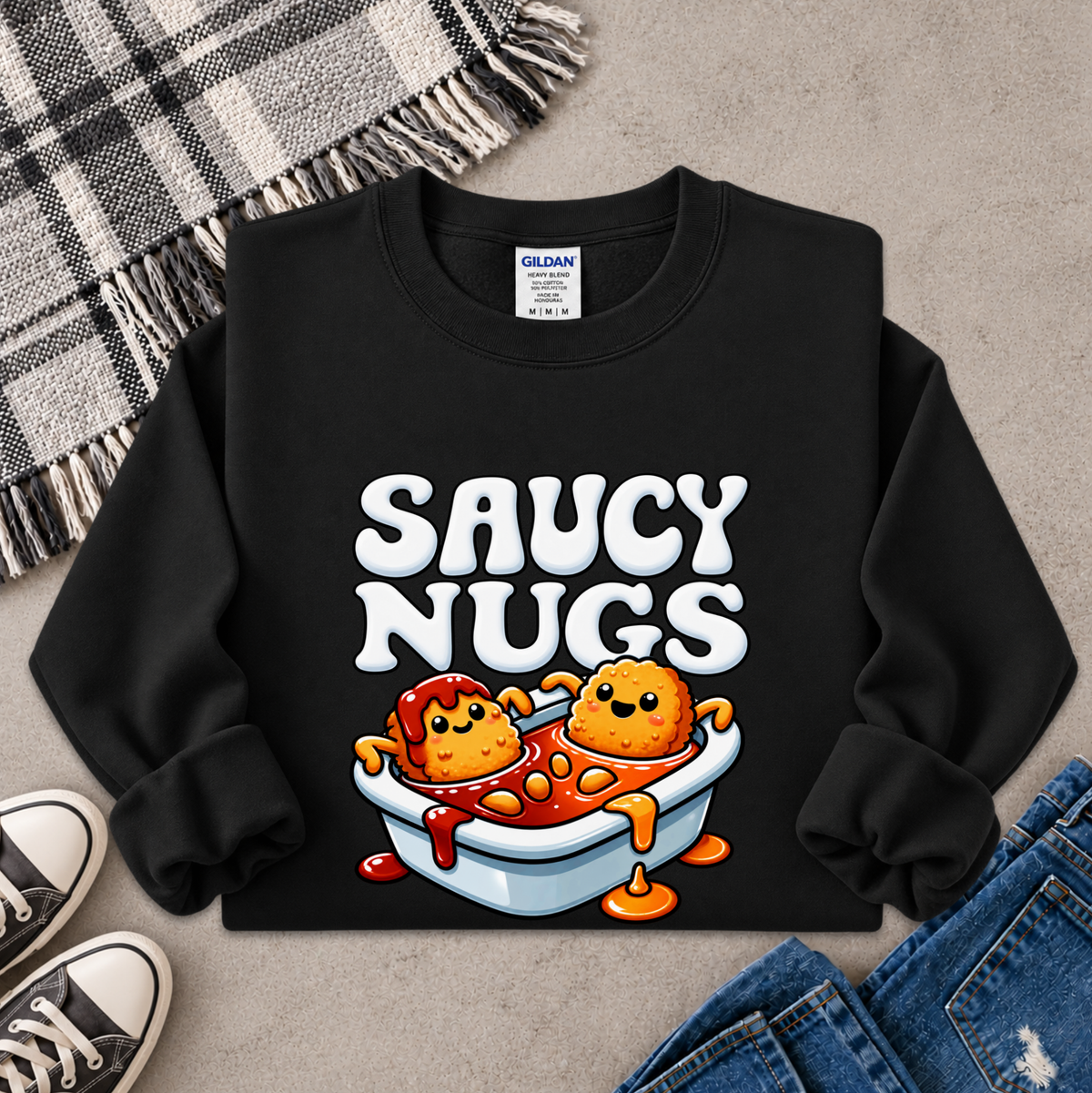 A black crewneck sweatshirt with a colorful graphic of two cartoon meatballs in a bowl of sauce under the words 'SAUCY NUGS'. The sweatshirt is styled in a flat lay with a plaid blanket, sneakers, and jeans on a gray concrete background.