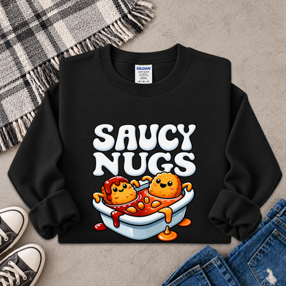 A black crewneck sweatshirt with a colorful graphic of two cartoon meatballs in a bowl of sauce under the words 'SAUCY NUGS'. The sweatshirt is styled in a flat lay with a plaid blanket, sneakers, and jeans on a gray concrete background.