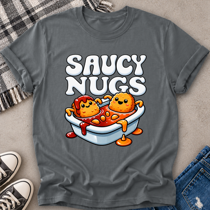 A dark gray graphic t-shirt laid flat on a neutral surface, featuring a cartoon design of two nuggets in a bathtub filled with sauce, with the text 'SAUCY NUGS' above. Styled flat-lay props include a plaid blanket, jeans, and sneakers.
