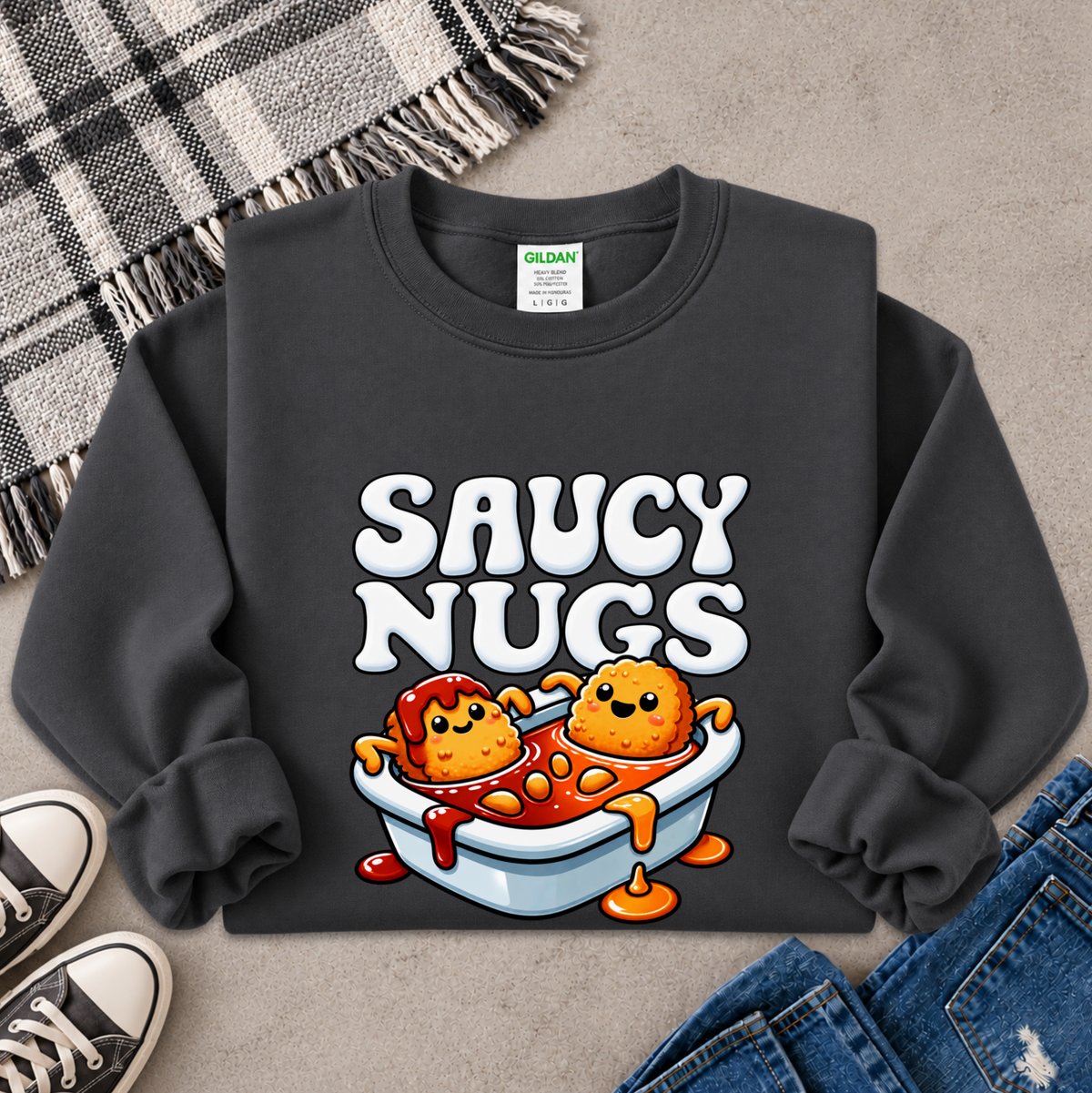 A dark charcoal gray sweatshirt laid flat on a textured gray surface, featuring a colorful graphic of two cartoon chicken nuggets in a bowl with the text 'SAUCY NUGS' in white. A Gildan brand tag is visible inside the collar.