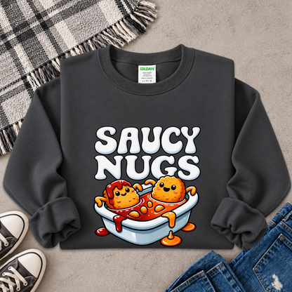 A dark charcoal gray sweatshirt laid flat on a textured gray surface, featuring a colorful graphic of two cartoon chicken nuggets in a bowl with the text 'SAUCY NUGS' in white. A Gildan brand tag is visible inside the collar.