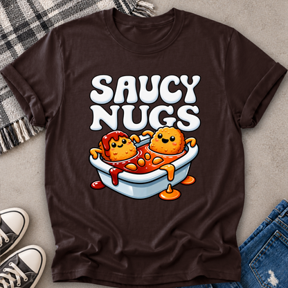 A dark brown graphic t-shirt laid flat on a gray textured surface with a plaid blanket, jeans, and sneakers as styling props. The shirt features a cartoon graphic of two orange nuggets sitting in a white bathtub filled with red sauce, with the text 'SAUCY NUGGTS' in large white letters above.