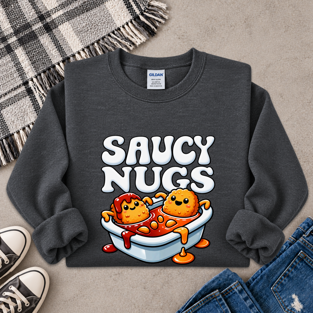 A dark gray crewneck sweatshirt featuring a cartoon graphic of two smiling orange nuggets in a white bowl of red sauce, with the text 'SAUCY NUGGS' above them. The sweatshirt is styled in a flat lay on a textured gray surface with a plaid blanket, jeans, and sneakers.
