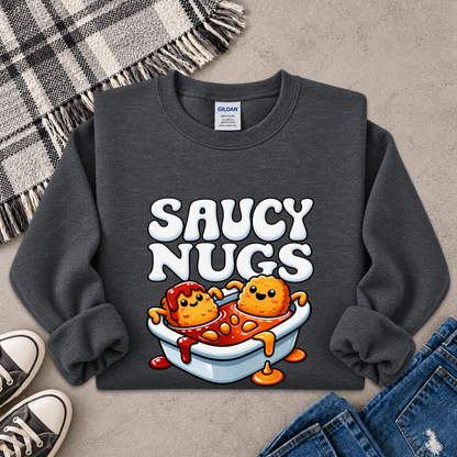 A dark gray crewneck sweatshirt featuring a cartoon graphic of two smiling orange nuggets in a white bowl of red sauce, with the text 'SAUCY NUGGS' above them. The sweatshirt is styled in a flat lay on a textured gray surface with a plaid blanket, jeans, and sneakers.