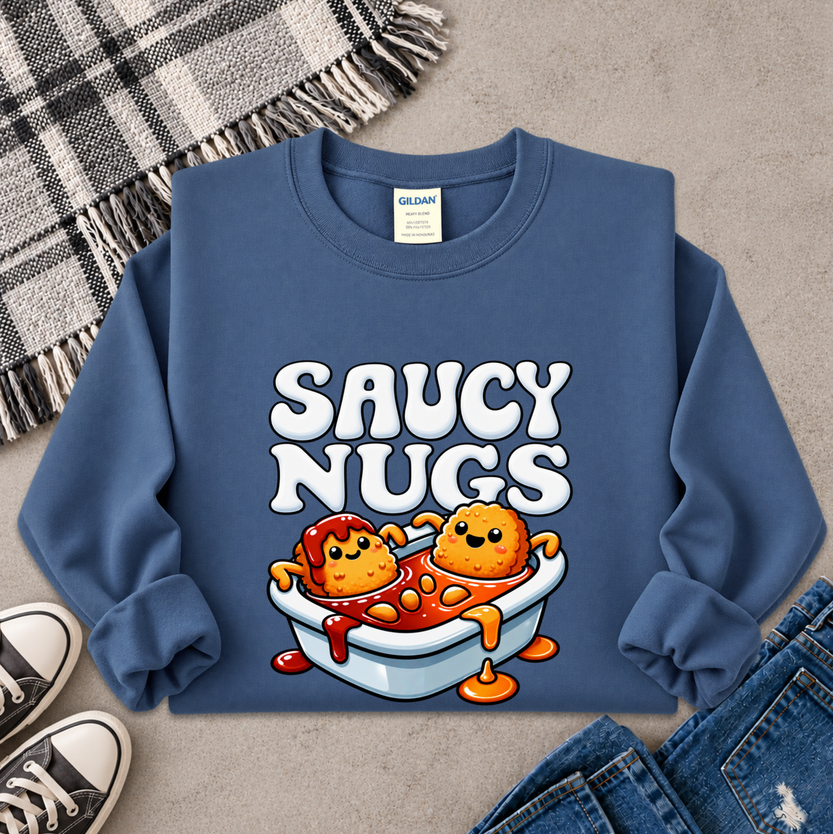 A blue crewneck sweatshirt laid flat on a gray surface, featuring a cartoon graphic of two orange nuggets in a white tub with red sauce and the text 'SAUCY NUGS'. A Gildan tag is visible inside the collar.
