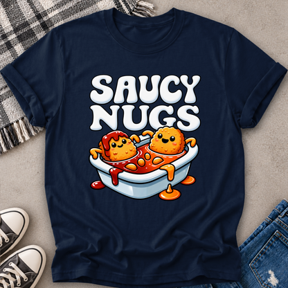 A navy blue graphic t-shirt laid flat on a neutral surface, featuring a cartoon design of two nuggets in a bathtub filled with sauce, with the text 'SAUCY NUGS' above. Styled flat-lay props include a plaid blanket, jeans, and sneakers.