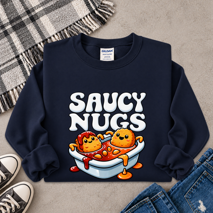 A navy blue crewneck sweatshirt with a cartoon graphic of two orange nuggets in a tub of red sauce, printed with the text 'SAUCY NUGS' in white. The sweatshirt is displayed in a flat-lay arrangement on a gray concrete floor with a plaid blanket, sneakers, and jeans.