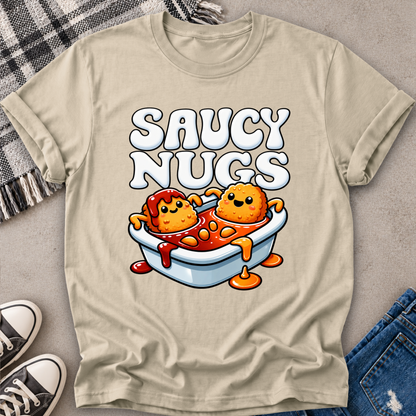 A beige graphic t-shirt laid flat on a gray surface with a plaid blanket, sneakers, and jeans around it. The shirt features a cartoon graphic of two orange nuggets sitting in a light blue bathtub filled with red sauce, with the text 'SAUCY NUGGS' above them.