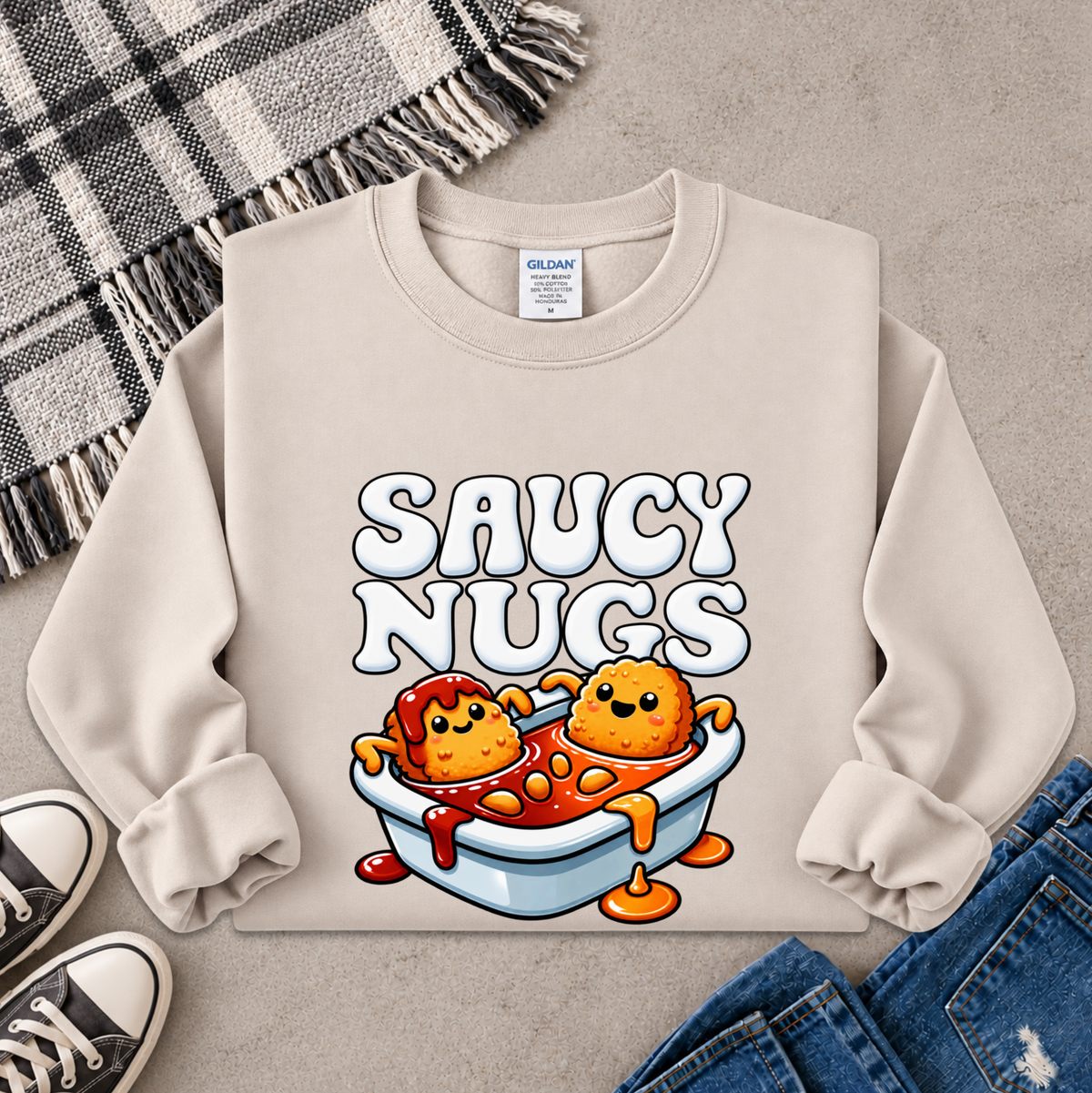 A beige crewneck sweatshirt laid flat on a gray surface, featuring a colorful graphic of two cartoon chicken nuggets in a bowl of orange sauce with the text 'SAUCY NUGS'. The sweatshirt is styled with a plaid blanket, black sneakers, and blue jeans.