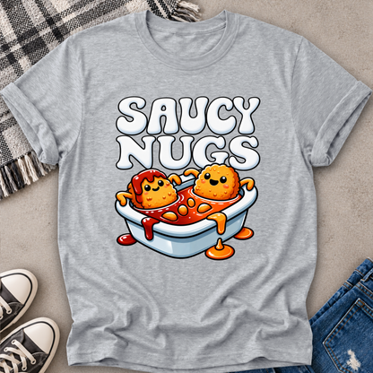 A heather gray short-sleeve t-shirt laid flat on a neutral surface, featuring a colorful cartoon graphic of two nuggets in a bathtub with the text 'SAUCY NUGS'. The shirt is styled with a plaid blanket, black sneakers, and blue jeans.