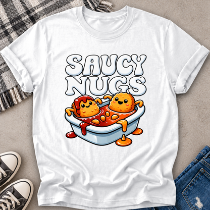 A white short-sleeve graphic T-shirt laid flat on a light gray surface, styled with a plaid blanket, sneakers, and jeans. The shirt features a cartoon graphic of two orange nuggets sitting in a bathtub filled with red sauce, with the text 'SAUCY NUGGTS' above them.