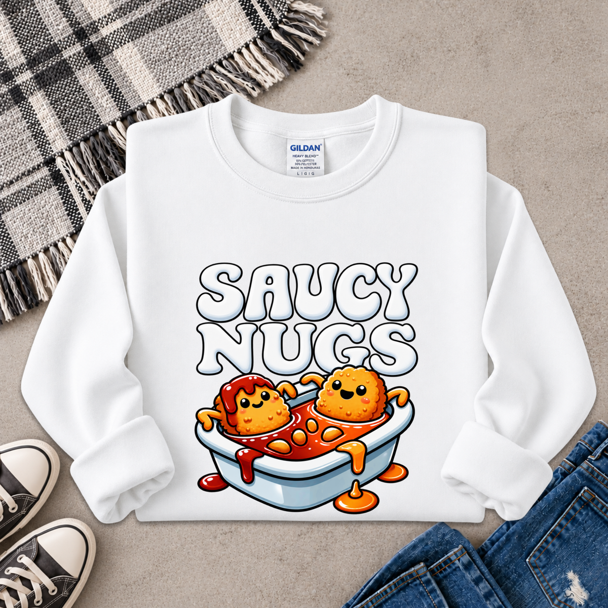 A white long-sleeve crewneck sweatshirt with a colorful graphic of two cartoon chicken nuggets in a bathtub labeled 'SAUCY NUGS'. The sweatshirt is styled in a flat-lay arrangement on a gray textured surface with a plaid blanket, black sneakers, and blue jeans.