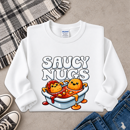 A white long-sleeve crewneck sweatshirt with a colorful graphic of two cartoon chicken nuggets in a bathtub labeled 'SAUCY NUGS'. The sweatshirt is styled in a flat-lay arrangement on a gray textured surface with a plaid blanket, black sneakers, and blue jeans.