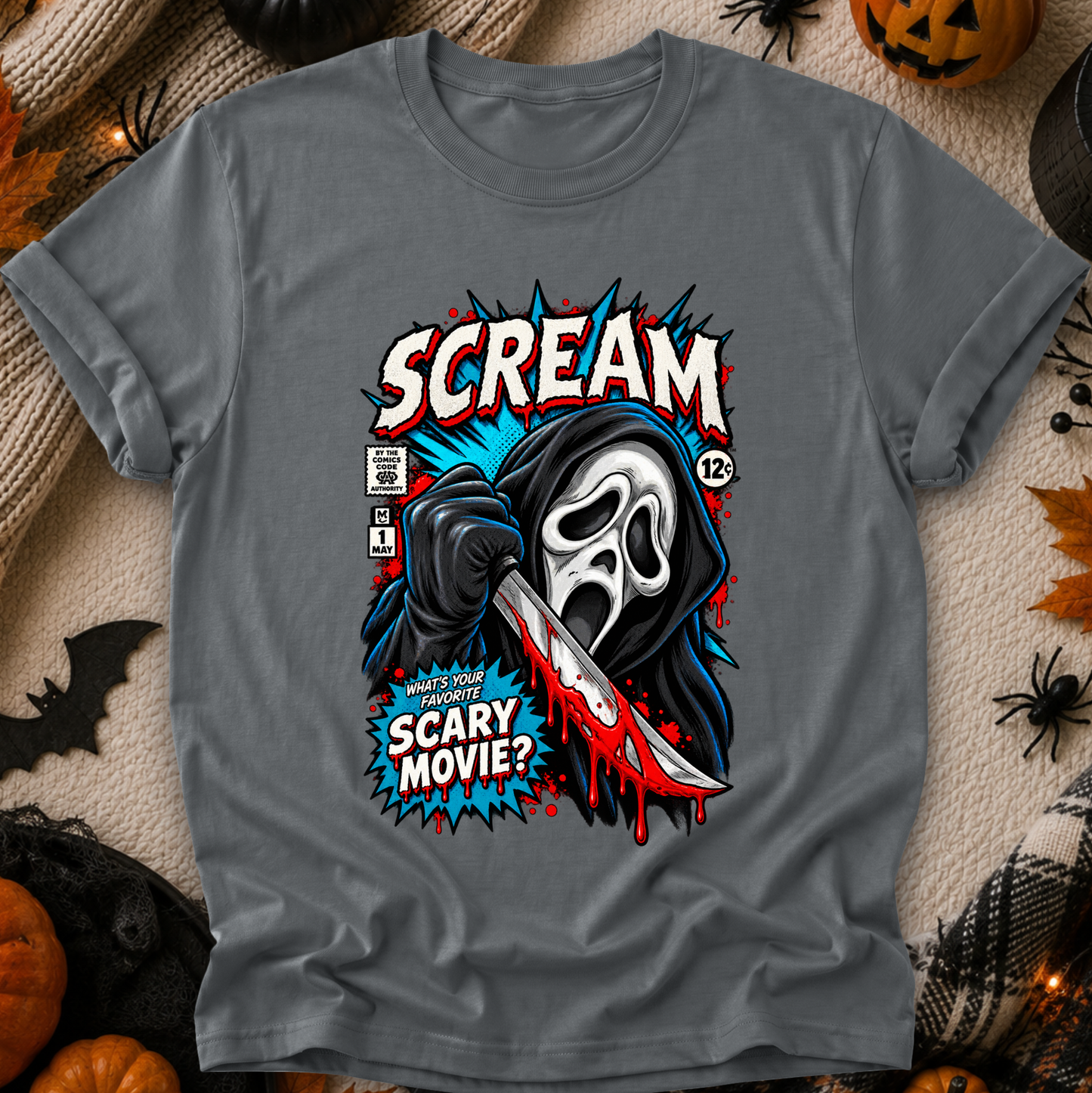 A gray short-sleeve graphic T-shirt laid flat on a cozy beige fabric background with Halloween props. The shirt features a large comic-style print of the Scream character holding a knife, with the words “SCREAM” and “SCARY MOVIE?” in bold red, white, blue, and black.