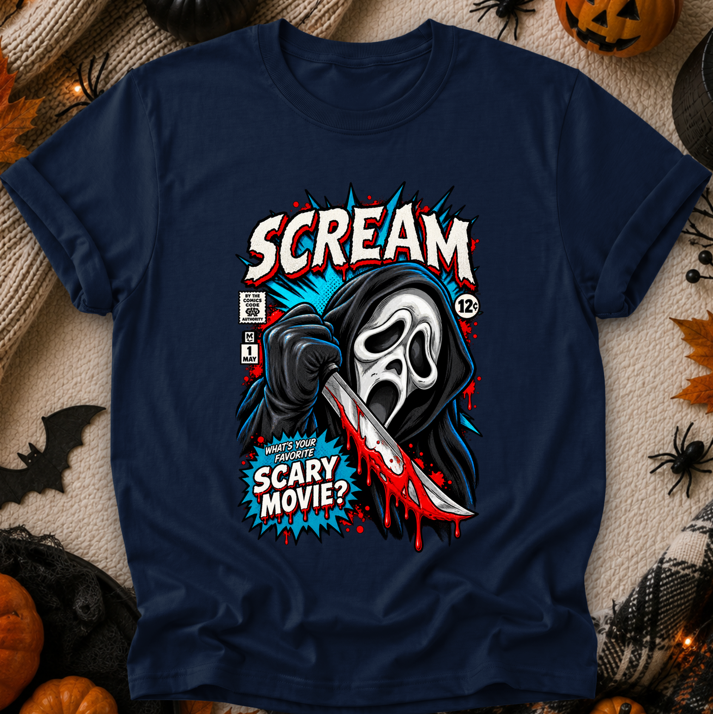 A navy blue graphic t-shirt featuring a stylized comic-book illustration of the Scream character holding a bloody knife, with the text 'SCREAM' and 'WHATS YOUR FAVORITE SCARY MOVIE?' in red, white, and blue. The shirt is displayed flat on a cozy beige knit blanket surrounded by Halloween decorations including pumpkins, a spider, and a bat.