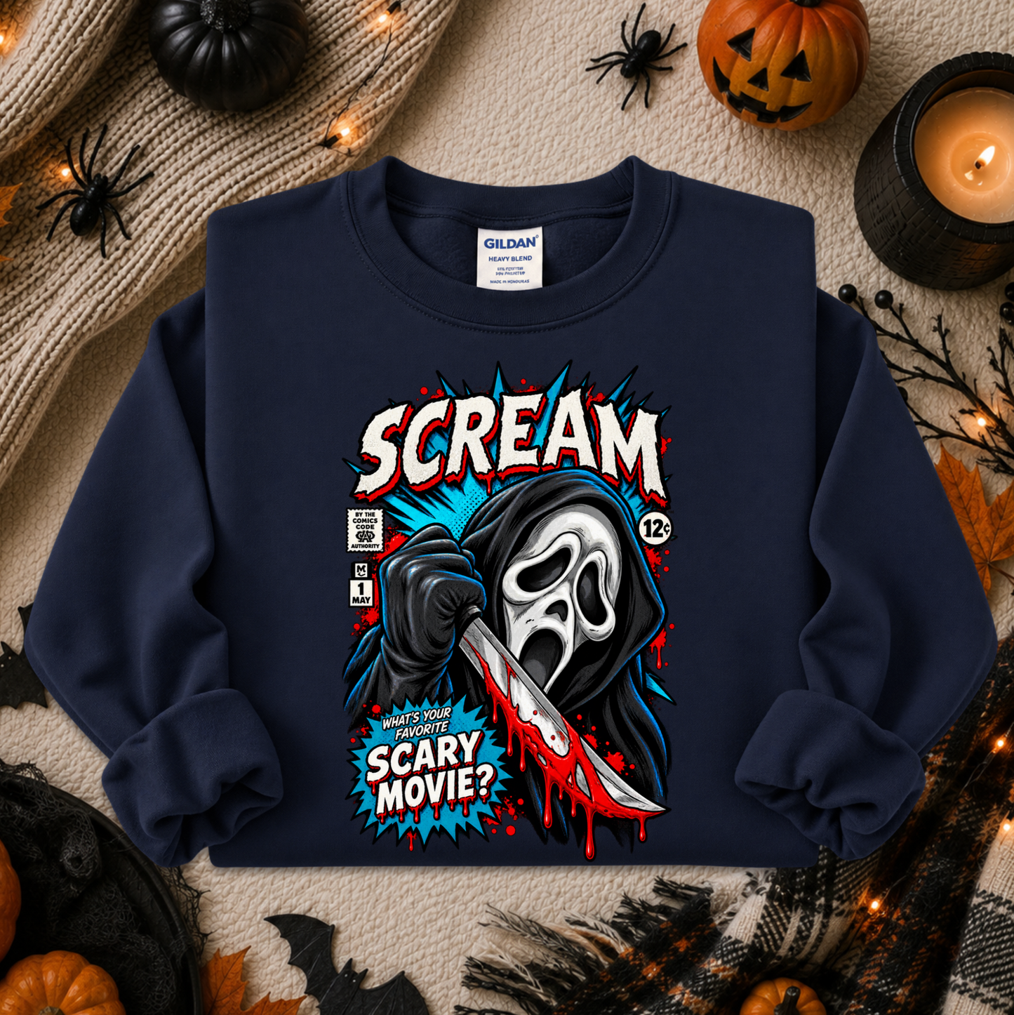 A navy blue crewneck sweatshirt with a comic-book style graphic of Ghostface holding a bloody knife, with the text 'SCREAM' and 'SCARY MOVIE?'. The sweatshirt is displayed in a flat-lay arrangement with Halloween props including pumpkins, a candle, and spider decorations on a beige textured blanket.