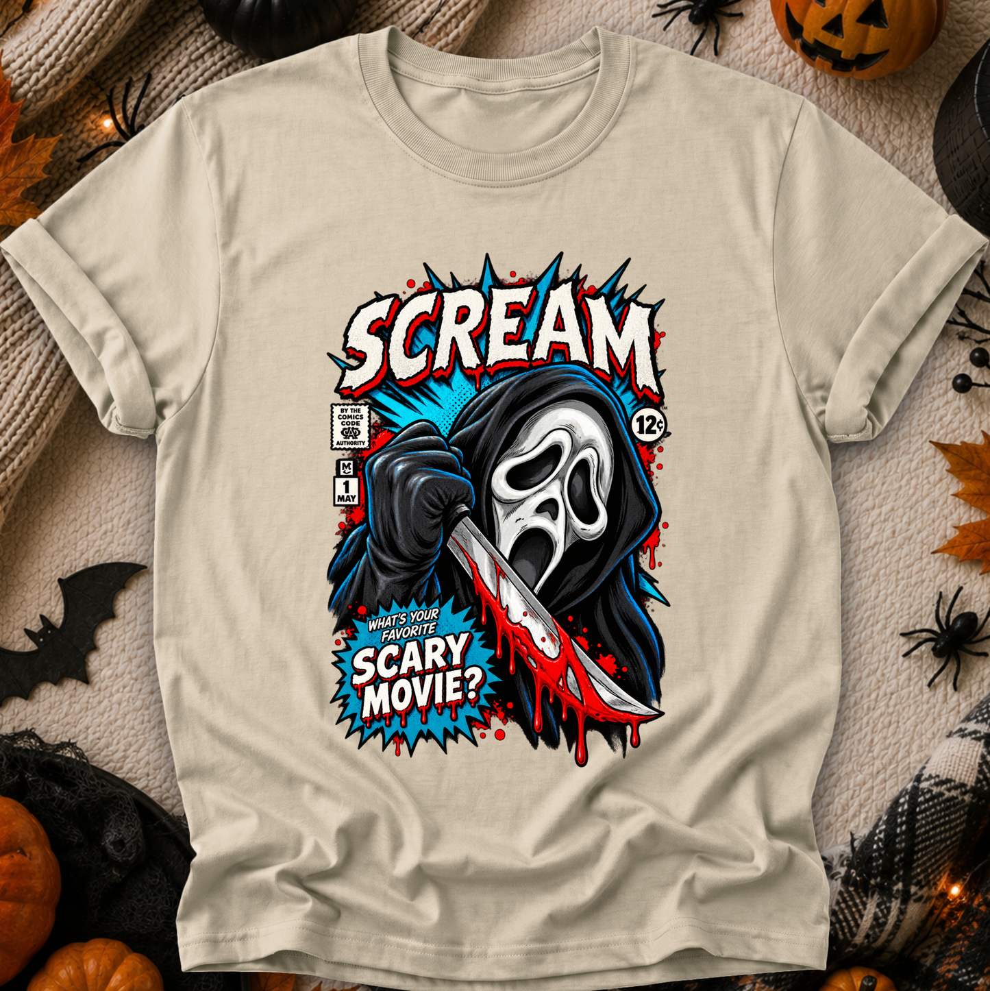 A beige graphic t-shirt featuring a horror movie design inspired by the Scream franchise, styled in a flat-lay with Halloween props like pumpkins, a spider, and a bat.