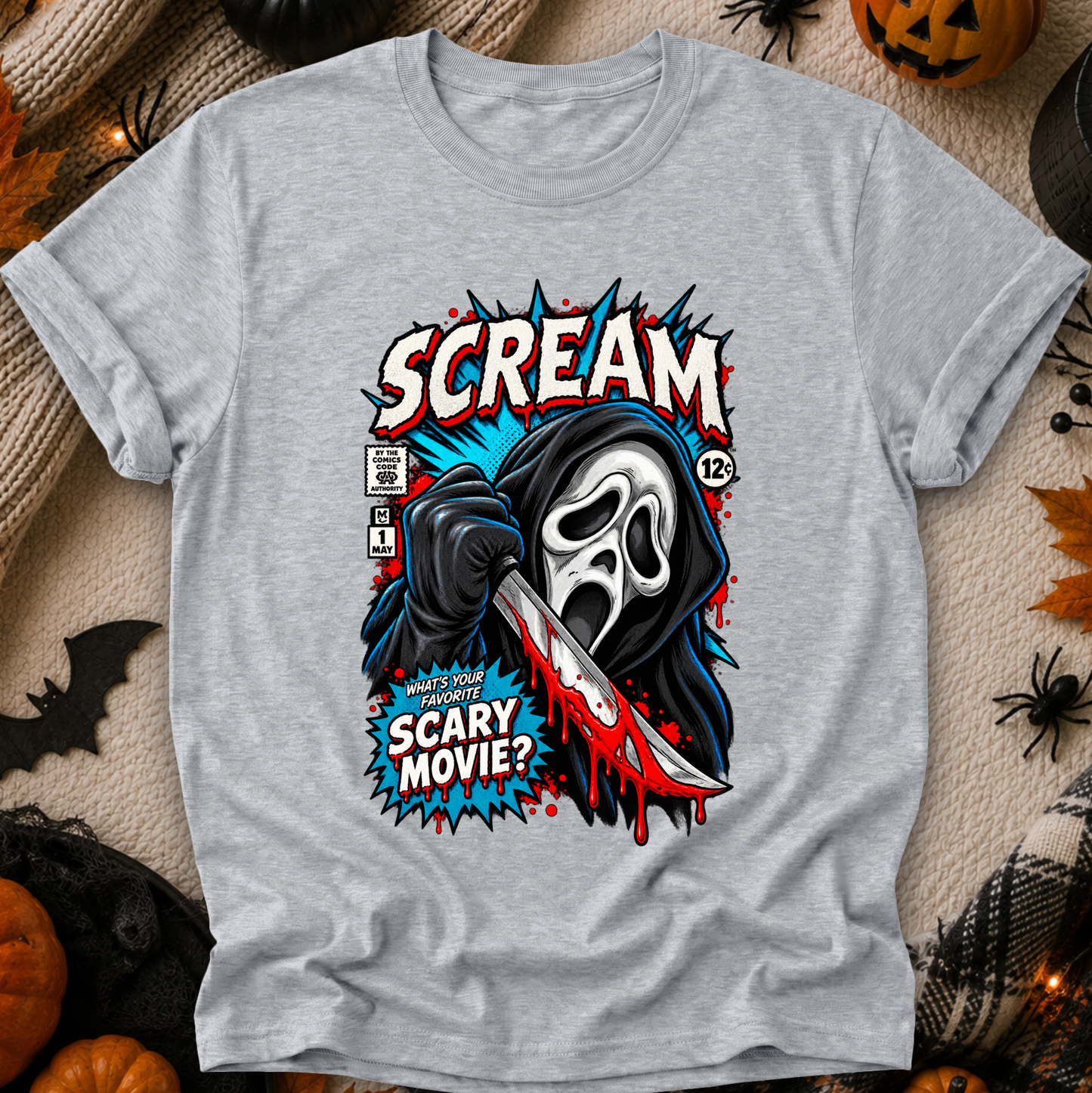 A heather gray short-sleeve t-shirt with a graphic print of the Ghostface character from Scream holding a bloody knife, surrounded by comic-style text and Halloween-themed props like pumpkins, spider decorations, and a black blanket.