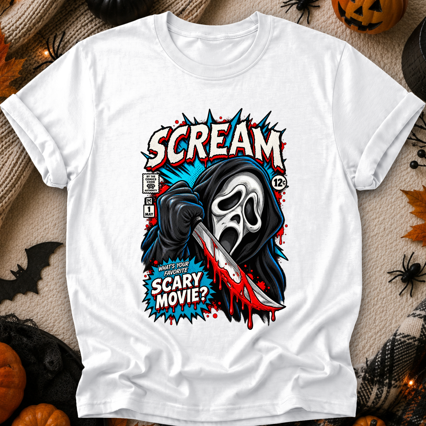 A white short-sleeve graphic T-shirt laid flat on a cozy beige fabric background with Halloween props. The shirt features a large front print of the Ghostface character holding a bloody knife, with bold text reading 'SCREAM' and 'SCARY MOVIE?' plus comic-style panel details.