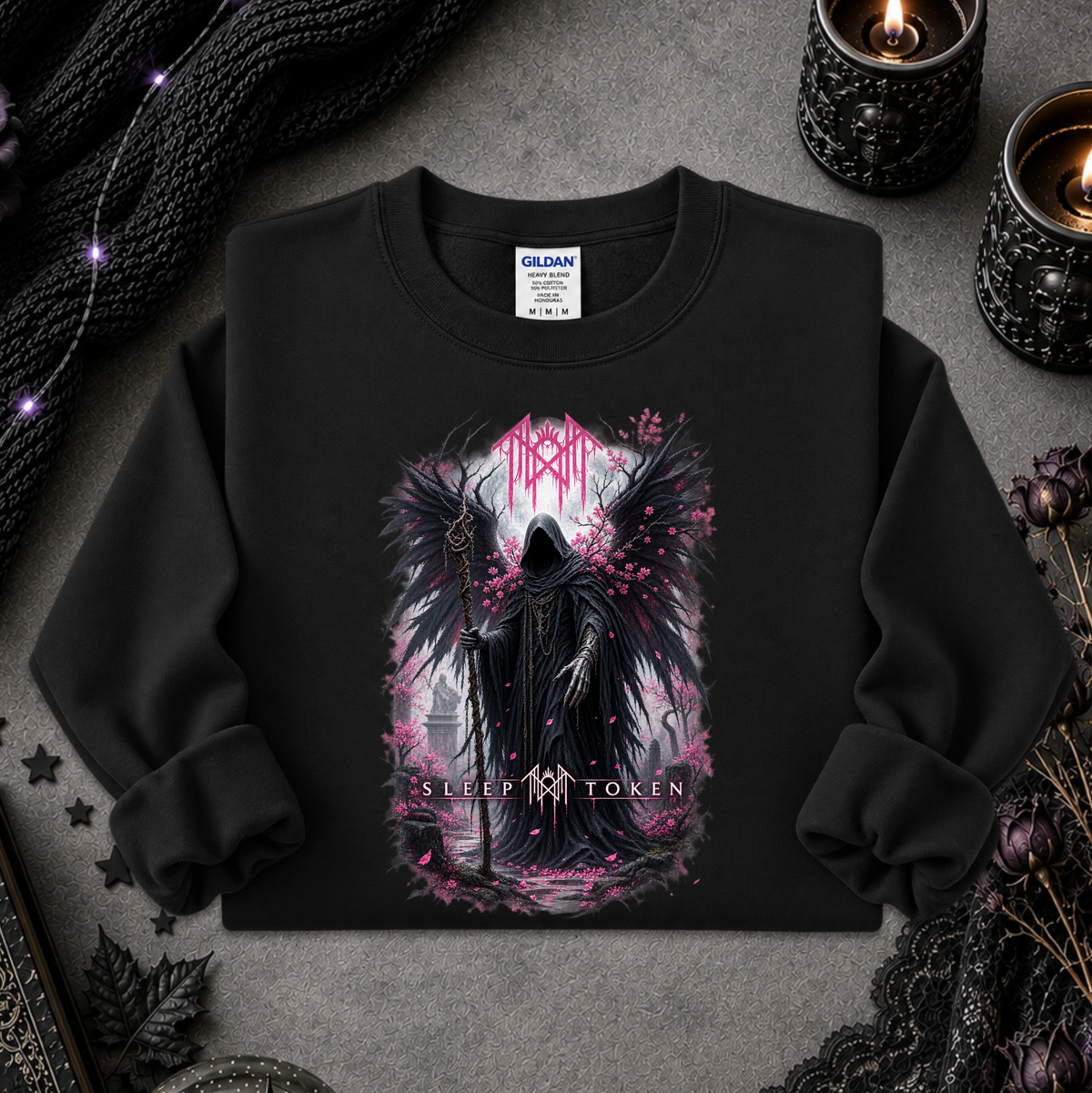 A black crewneck sweatshirt with a gothic graphic of a dark angel figure and pink accents, styled flat-lay on a gray textured surface with candles, lace, and a knit blanket.