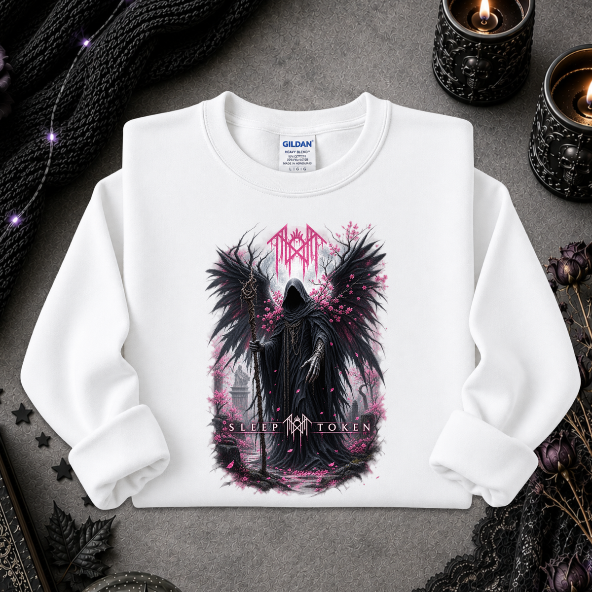 A white long-sleeve crewneck sweatshirt with a gothic graphic of a winged dark figure and pink text, laid flat on a gray surface with candles and black lace props.
