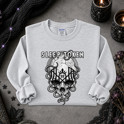 A light gray crewneck sweatshirt with a black graphic of a skull, snakes, and the words 'SLEEP TOKEN' on the front, laid flat on a gray surface with candles and dark fabric props around it.