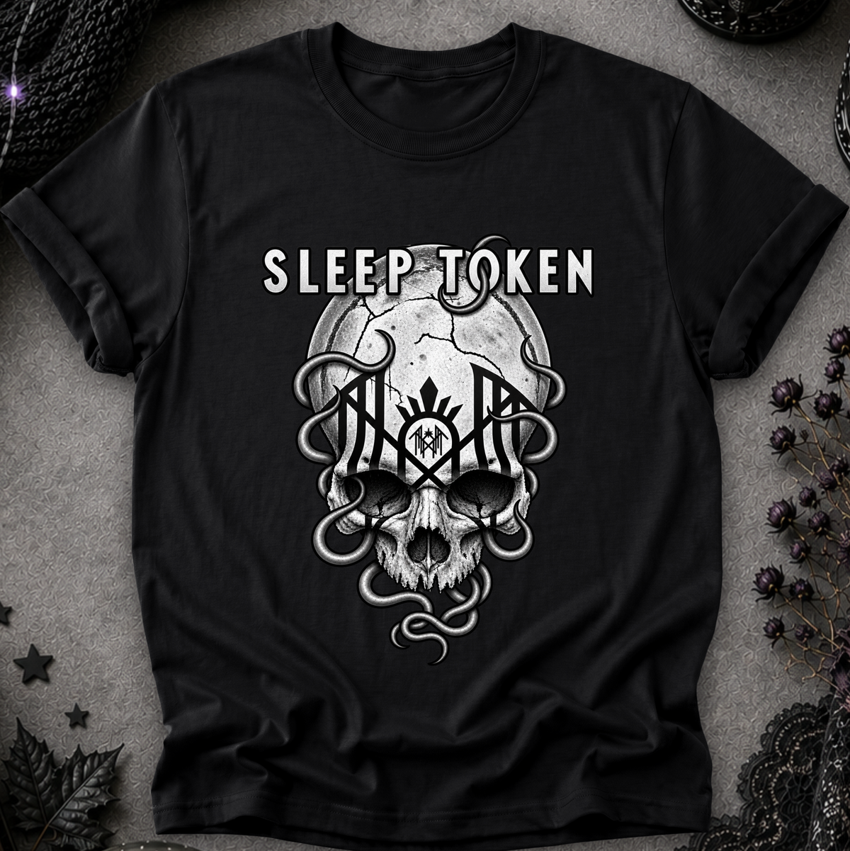 A black short-sleeve graphic t-shirt featuring a white and gray gothic design with the text 'SLEEP TOKEN' above a skull and snake motif, laid flat on a textured gray background with dark decorative props.