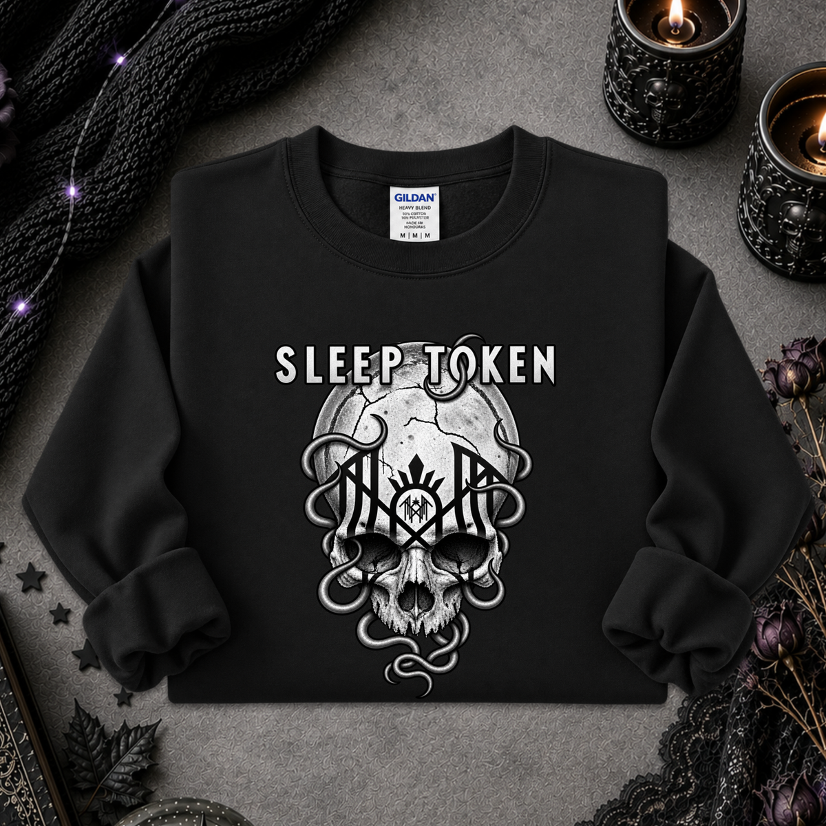 A black crewneck sweatshirt with a white graphic of a skull and the words 'SLEEP TOKEN' printed on the front, styled in a flat-lay with candles, lace, and a knit scarf on a gray textured background.