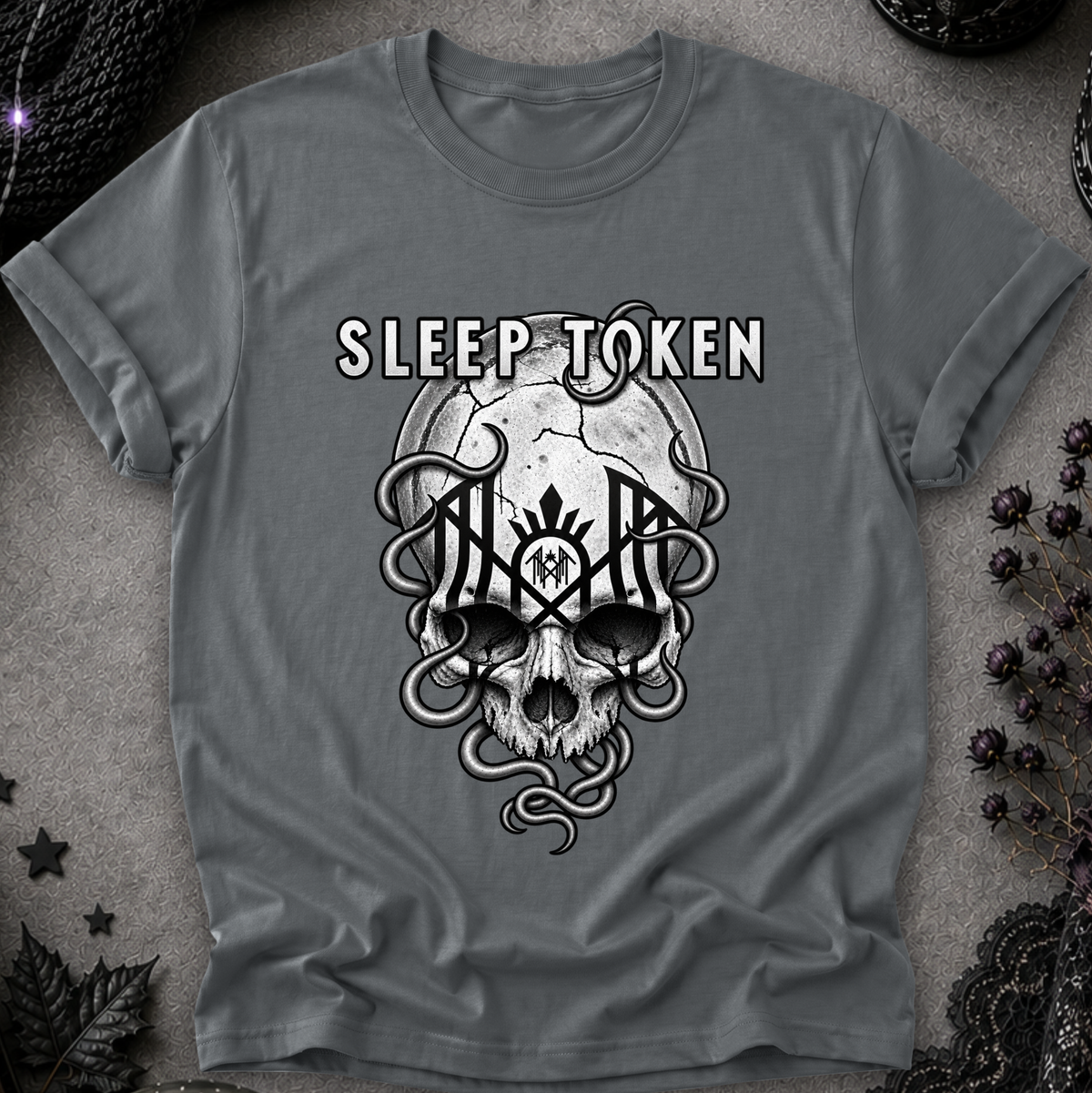A gray short-sleeve t-shirt with a black and white graphic of a skull, snakes, and the text 'SLEEP TOKEN' laid flat on a textured gray background with dark decorative props.