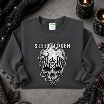 A dark gray crewneck sweatshirt featuring a gothic graphic of a skull with snakes and the text 'SLEEP TOKEN' above it. The sweatshirt is laid flat on a textured gray surface surrounded by spooky props like black lace, dried flowers, and lit candles.
