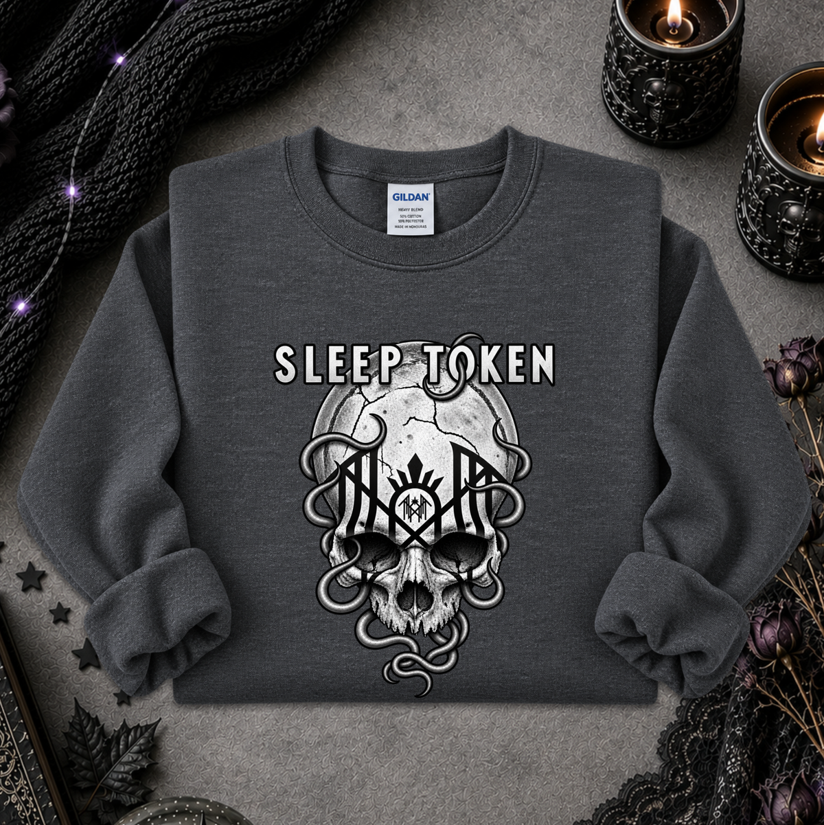 A dark gray crewneck sweatshirt featuring a gothic graphic of a skull with snakes and the words 'SLEEP TOKEN' above it. The sweatshirt is laid flat on a textured gray surface surrounded by decorative props including black lace, dried flowers, and lit candles.