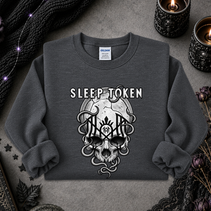 A dark gray crewneck sweatshirt featuring a gothic graphic of a skull with snakes and the words 'SLEEP TOKEN' above it. The sweatshirt is laid flat on a textured gray surface surrounded by decorative props including black lace, dried flowers, and lit candles.