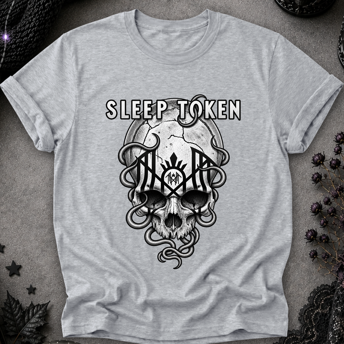 A light heather gray t-shirt featuring a black graphic of a skull with snakes and the words 'SLEEP TOKEN' above it, laid flat on a textured gray background with dark gothic props.