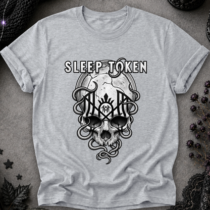 A light heather gray t-shirt featuring a black graphic of a skull with snakes and the words 'SLEEP TOKEN' above it, laid flat on a textured gray background with dark gothic props.