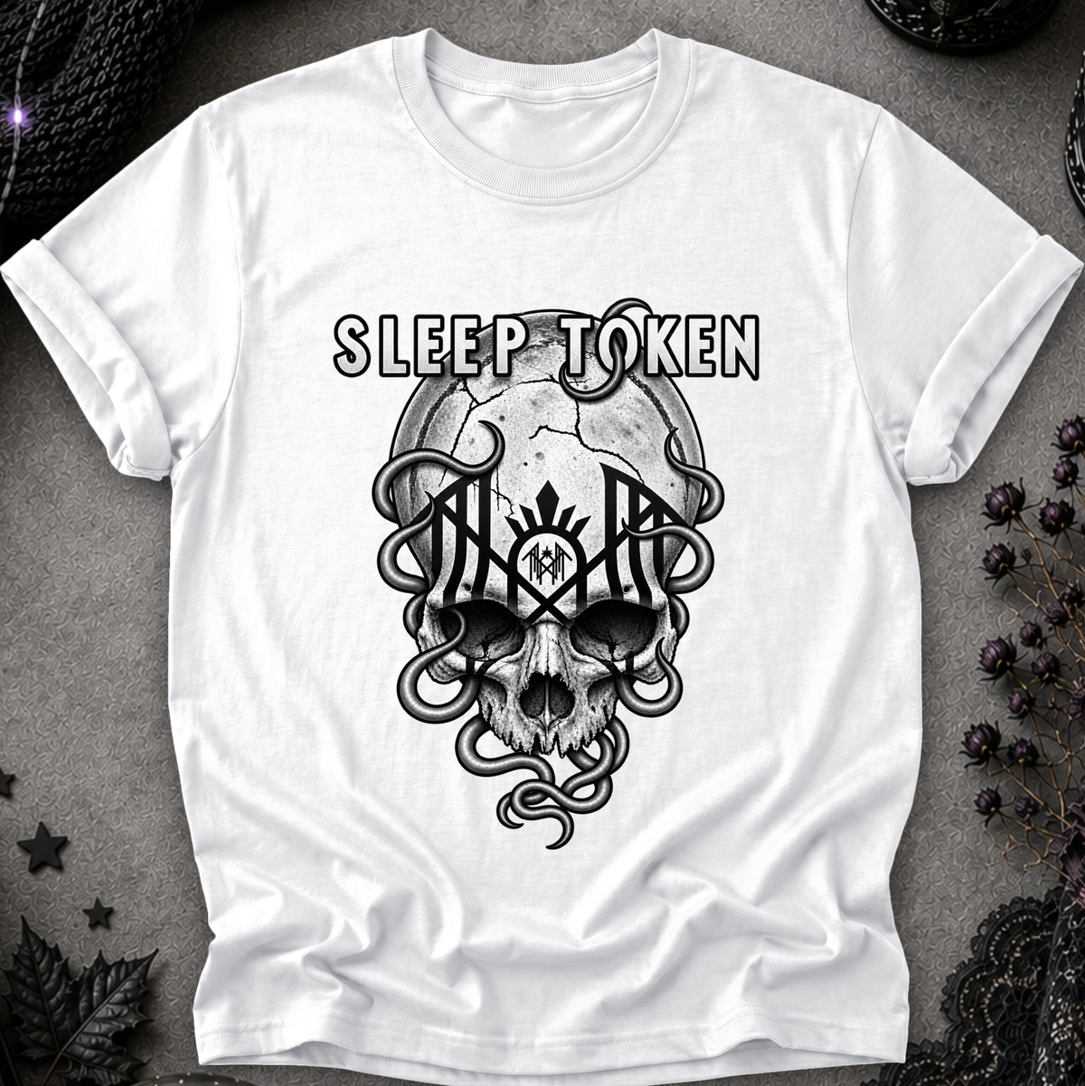 A white short-sleeve crewneck t-shirt featuring a black graphic print of a skull, snakes, and the text 'SLEEP TOKEN' on the front, laid flat on a textured gray background with gothic decorative props.