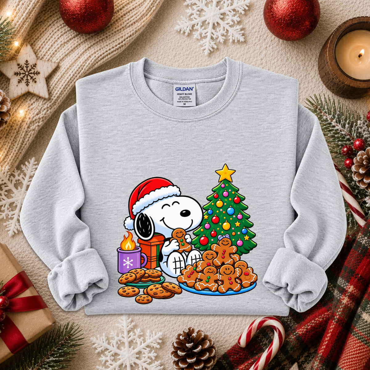 A light gray long-sleeve crewneck sweatshirt featuring a colorful graphic of Snoopy and Woodstock by Christmas, styled in a cozy holiday flat-lay with a knit blanket, red ornaments, a candle, and gingerbread cookies.