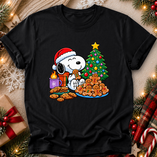 A black short-sleeve crewneck t-shirt featuring a festive Christmas graphic of Snoopy wearing a Santa hat and eating cookies next to a decorated tree and presents, displayed on a cozy holiday-themed background with pine branches, lights, and candy canes.