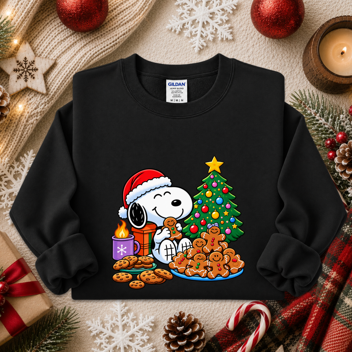 A black crewneck sweatshirt featuring a colorful graphic of Snoopy and Woodstock by Christmas, styled in a cozy holiday flat-lay with a knit blanket, ornaments, candy canes, and pinecones.