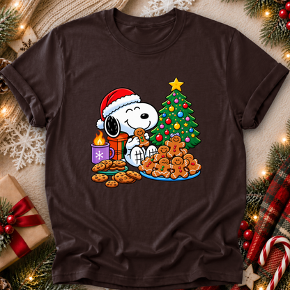 A dark brown short-sleeve crewneck t-shirt featuring a festive graphic of Snoopy wearing a Santa hat and eating a gingerbread cookie next to a decorated Christmas tree, cookies, and a gift box. The shirt is displayed in a flat-lay Christmas setting with pine branches, lights, and candy canes.