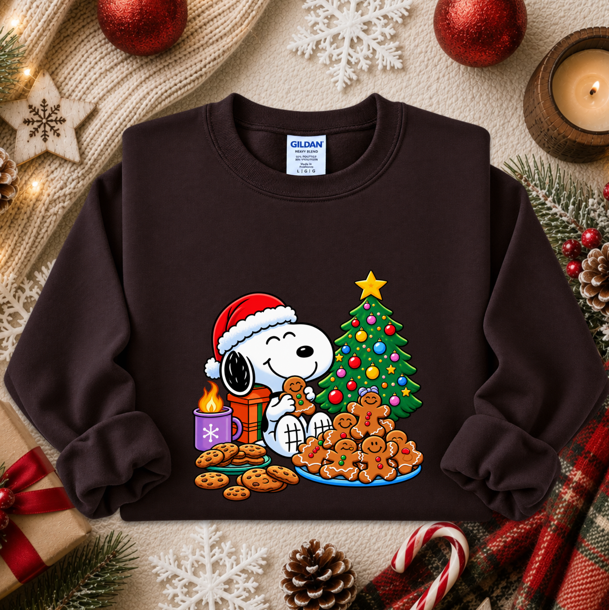 A dark brown crewneck sweatshirt featuring a colorful graphic of Snoopy and Woodstock by Peanuts decorating a Christmas tree and making gingerbread cookies. The sweatshirt is styled in a cozy holiday flat-lay with a knit blanket, candles, ornaments, and candy canes.