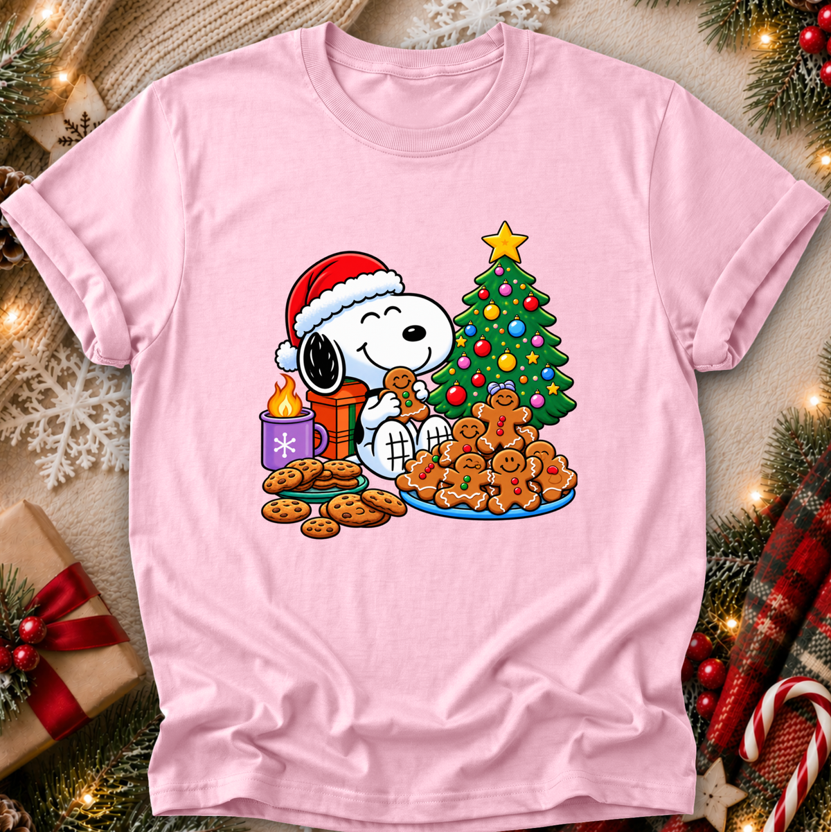 A light pink short-sleeve crewneck t-shirt featuring a festive Christmas graphic of Snoopy wearing a Santa hat and eating gingerbread cookies next to a decorated tree and gift box, displayed on a cozy holiday-themed background.