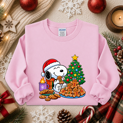 A pink long-sleeve crewneck sweatshirt featuring a colorful graphic of Snoopy and Woodstock by a Christmas tree, displayed in a cozy holiday flat-lay setting with a knit scarf, candle, ornaments, and candy canes.